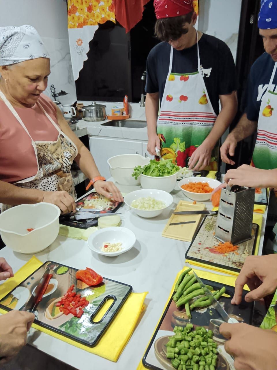 Group of people cooking and chopping vegetables in a kitchen, wearing colorful aprons and bandanas.