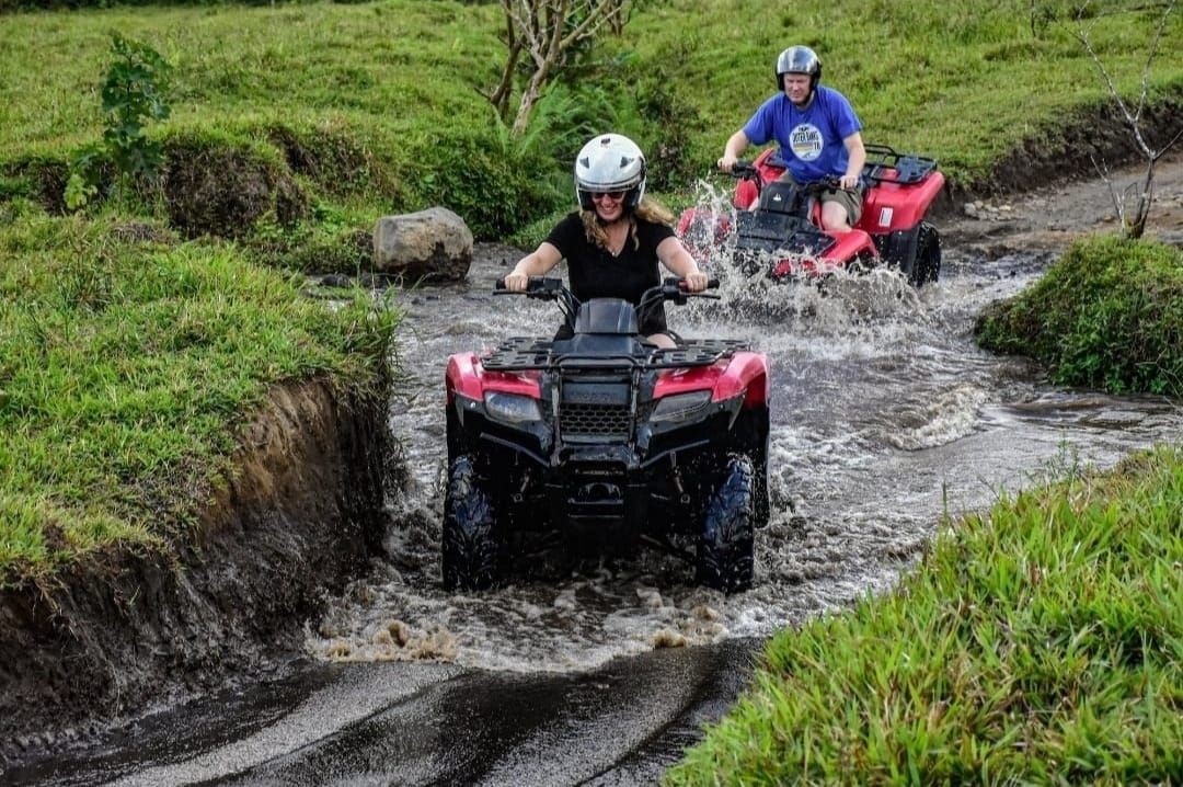 Two people riding red ATVs through a shallow stream in a grassy area.
